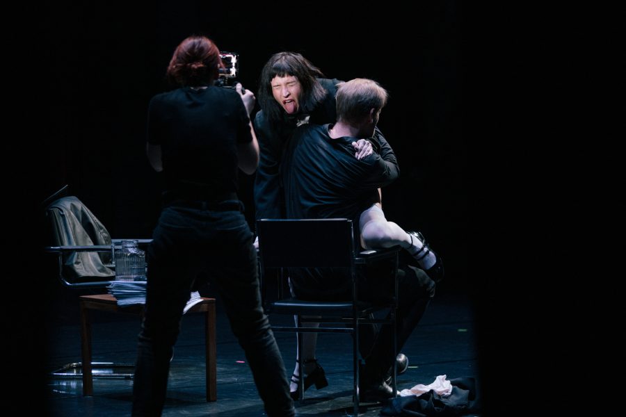A person films two actors onstage; one actor pulls a silly face while sitting on the other’s lap.