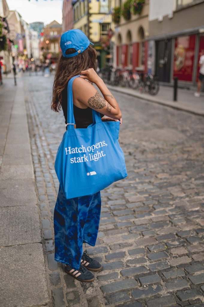 Woman in blue hat, trousers, and tote bag with text Hatch opens, stark light. standing on cobbled street.