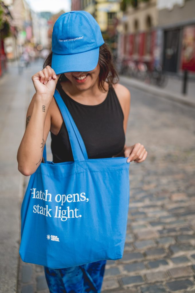 Smiling woman in blue cap and tote bag with text, walking on a cobbled street.