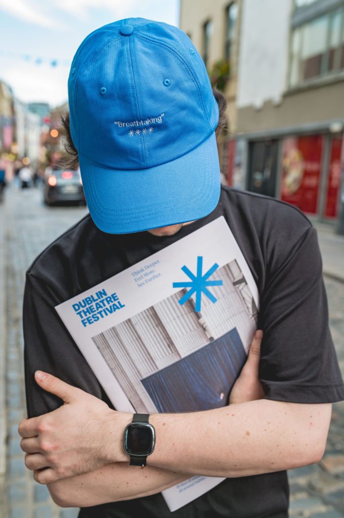 Person in a blue cap and black shirt hugging a Dublin Theatre Festival programme on a cobbled street.