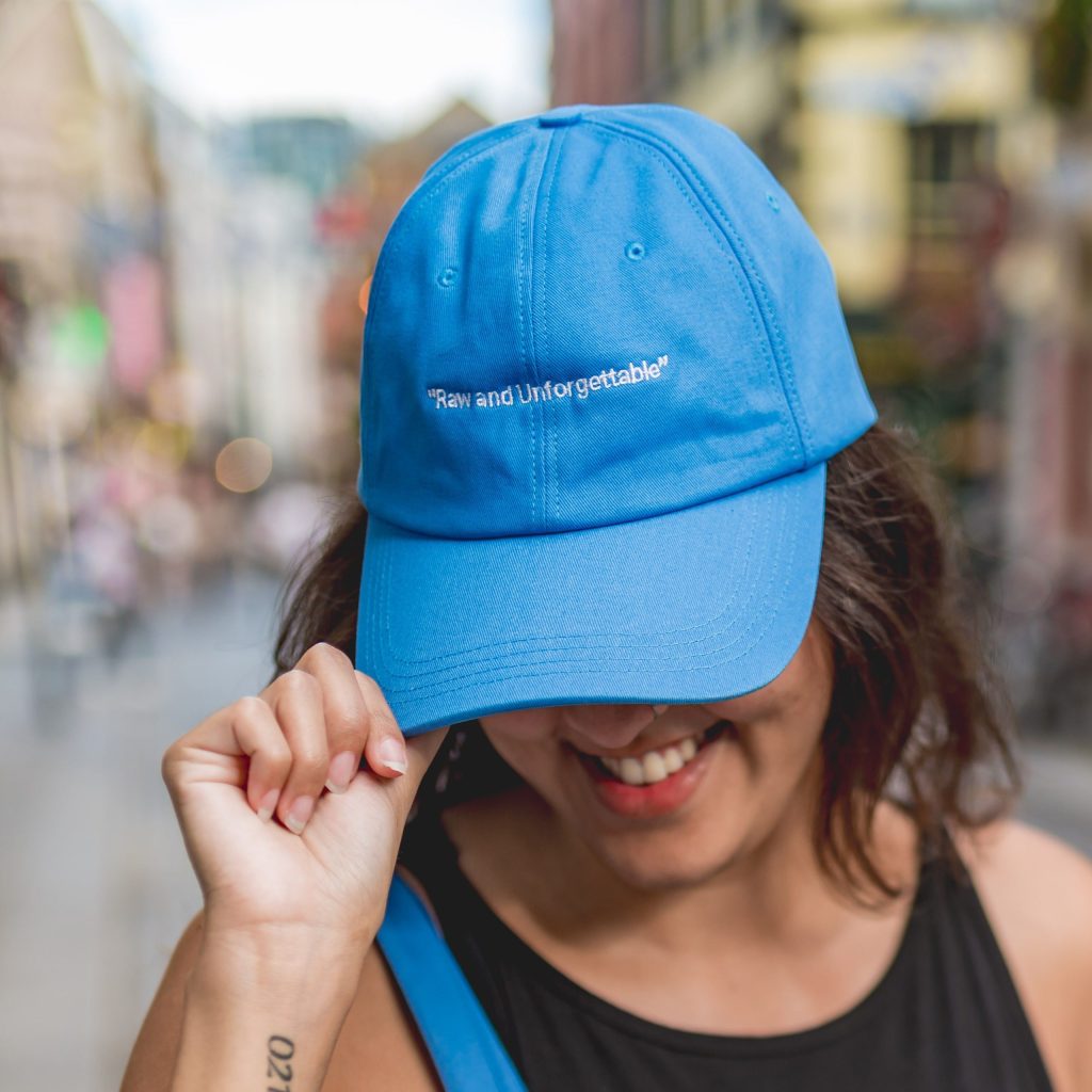 Smiling person in a blue cap that says Raw and Unforgettable, holding the peak, outdoors on a city street.