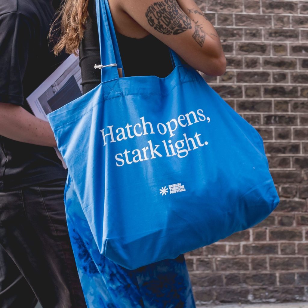 Person carrying a blue tote bag that reads Hatch opens, stark light. on a city street.